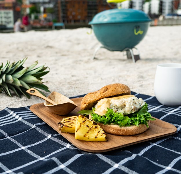 Strand med ananas i bakgrunnen, en hvit plastkopp, et blått håndkle med en fiskeburger oppå og grillet ananas. 