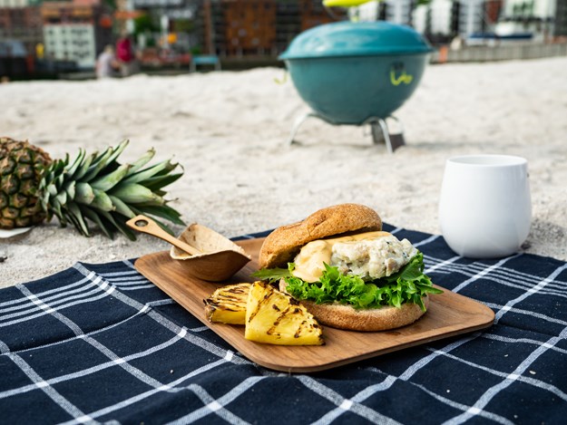 Strand med ananas i bakgrunnen, en hvit plastkopp, et blått håndkle med en fiskeburger oppå og grillet ananas. 