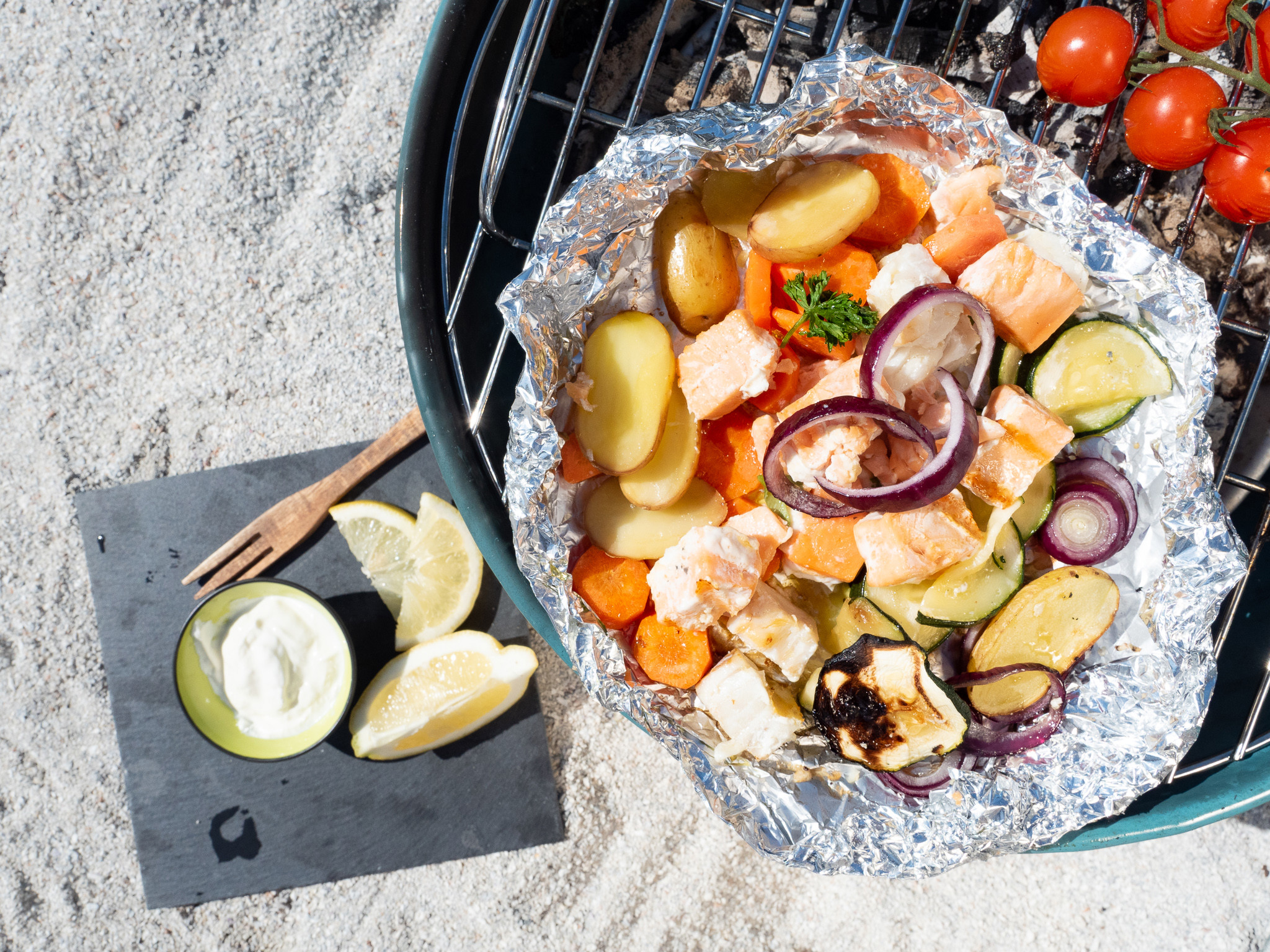 Strand med grill. Oppå grillen er aliminiumsfolie med grønnsaker og fisketerninger og tomater ved siden av.